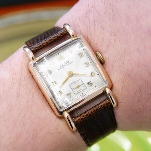 May include: A gold-toned wristwatch with a square face and a brown leather strap. The watch face has white Roman numeral hour markers and a small seconds dial at the 6 o'clock position. The watch is labelled "Venus" and "Swiss Made".