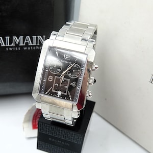 May include: A silver-toned Balmain Swiss watch with a rectangular face and a stainless steel band. The watch face is dark with subdials and the number 12 at the top. The watch is displayed on a black stand with the Balmain logo.