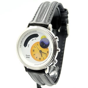 Könnte beinhalten: Eine silberfarbene Quarz-Armbanduhr mit schwarzem Lederarmband. Das Zifferblatt hat einen weißen Hintergrund mit einem gelb-orangenen Zifferblatt, einer blau-gelben Kugel und dem Text "THE THIRD MILLENNIUM CHALLENGE".