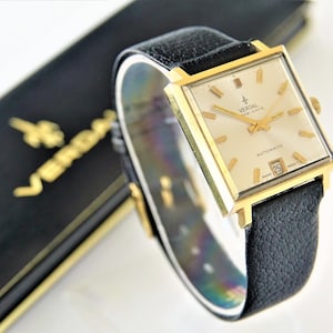 May include: A gold-toned, square-faced wristwatch with a black leather band. The watch face is cream-colored with gold hands and markers. The watch has the text "VERDAL" and "SWISS MADE" on the face. The watch is displayed next to a black box with gold lettering.