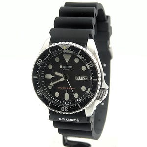 Può includere: Orologio subacqueo automatico Seiko nero con quadrante nero e datario. L'orologio ha una lunetta in acciaio inossidabile e un cinturino in gomma nero. Il quadrante ha indici e lancette bianche. Resistente all'acqua fino a 200m.