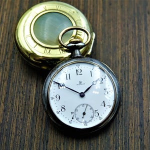 May include: A vintage pocket watch with a white face, black hands, and Roman numeral hour markers. The watch has a silver-colored case and a gold-colored top. The watch is open, revealing the time. The background is a dark wood surface.