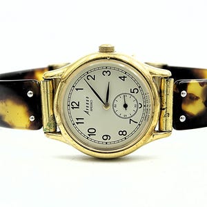 Peut inclure: Une montre Avenue Seiko dorée avec un cadran crème, des aiguilles noires et un petit cadran de secondes. La montre a un bracelet à motif écaille de tortue. Le cadran affiche les chiffres de 1 à 12.