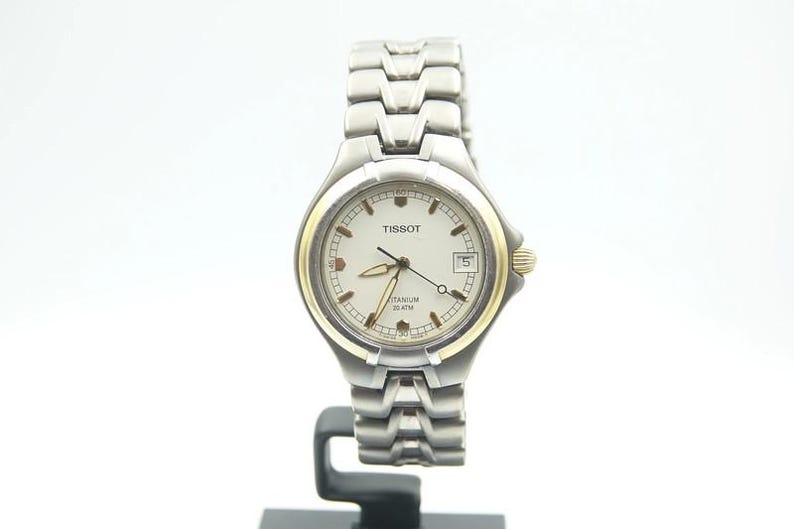 May include: A silver and gold Tissot wristwatch with a white face and a titanium band. The watch face has the words "Tissot" and "Titanium 100 meter" printed on it.