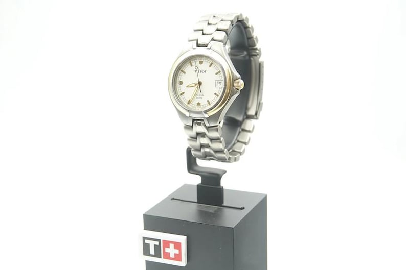 May include: A silver and gold wristwatch with a white face and a date window. The watch is on a black display stand with a T+ logo.