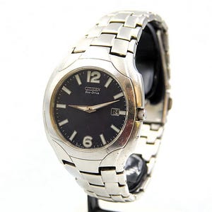 May include: A Citizen Eco-Drive wristwatch with a silver-toned stainless steel band and a black watch face. The watch features white hour markers, a date display at the 3 o'clock position, and luminous hands. The watch case is approximately 40mm in diameter.