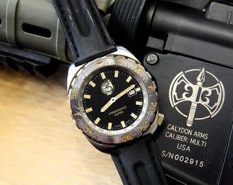 May include: A black and gold wristwatch with a black leather band. The watch face is black with gold accents and the text "STREMENTO ALPINI". The watch is next to a black firearm with the text "CALYDON ARMS".