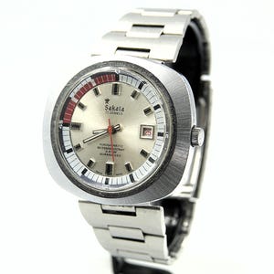 May include: A vintage Sakata wristwatch with a silver-colored face and a stainless steel band. The watch face features red and black accents, a date display, and the text "Sakata 17 Jewels". The watch has a rectangular case.