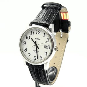 Könnte beinhalten: Eine silberne Timex-Uhr mit weißem Zifferblatt, schwarzen Ziffern und Datumsanzeige auf 3 Uhr. Die Uhr hat schwarze Zeiger und ein schwarzes Lederarmband. Die Uhr ist vor weißem Hintergrund.