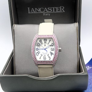 May include: A Lancaster Italy wristwatch with a tonneau-shaped silver case encrusted with pink stones. The white dial features large black Arabic numerals, blue hands, and the brand name. It has a light beige leather strap and is presented in a box.