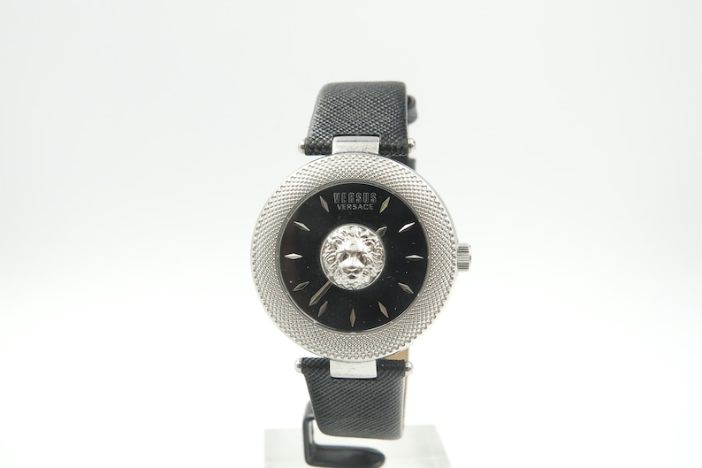 Puede incluir: Un reloj de pulsera Versus Versace con esfera negra, un emblema central de cabeza de le&oacute;n plateada y un bisel plateado texturizado. El reloj cuenta con una correa de cuero negro texturizado y herrajes plateados, con el nombre de la marca impreso.