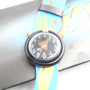 May include: A black wristwatch with a flame design on the strap and a black face with a flame graphic. The watch strap is blue with yellow and orange flames. The watch is on a grey surface.