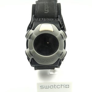 Może przedstawiać: Czarny i szary zegarek Swatch z czarnym paskiem z tkaniny. Tarcza zegarka jest szara z czarnym środkiem i białymi przyciskami. Na tarczy i pasku zegarka widnieje napis "Swatch".