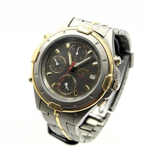 May include: A silver and gold-toned Racer chronograph watch with a dark gray face. The watch features three sub-dials, red hands, and a date display. The watch band is a combination of silver and gold-toned metal links.