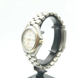 May include: A silver and gold wristwatch with a white face and a silver metal band. The watch is on a display stand.