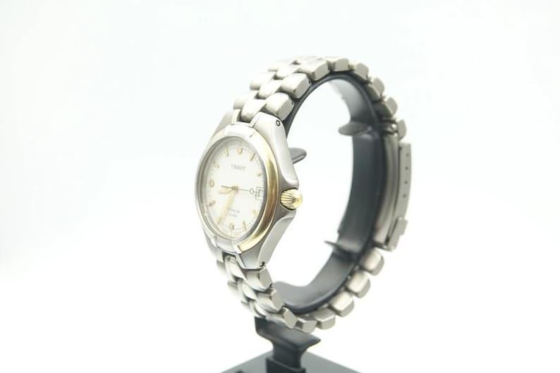 May include: A silver and gold wristwatch with a white face and a silver metal band. The watch is on a display stand.