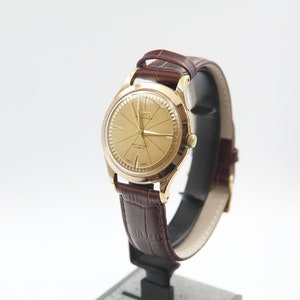 Może przedstawiać: Złoty zegarek na rękę z brązowym skórzanym paskiem. Tarcza zegarka ma wzór promienisty, a na niej nadrukowane są słowa "ZENO" i "Automatic".