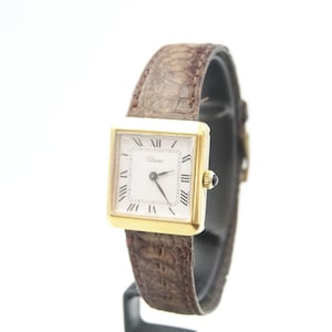 Op de afbeelding: Een goudkleurig horloge met een vierkante wijzerplaat en Romeinse cijfers. Het horloge heeft een witte wijzerplaat en een bruine leren band. Het horloge wordt gepresenteerd tegen een witte achtergrond.
