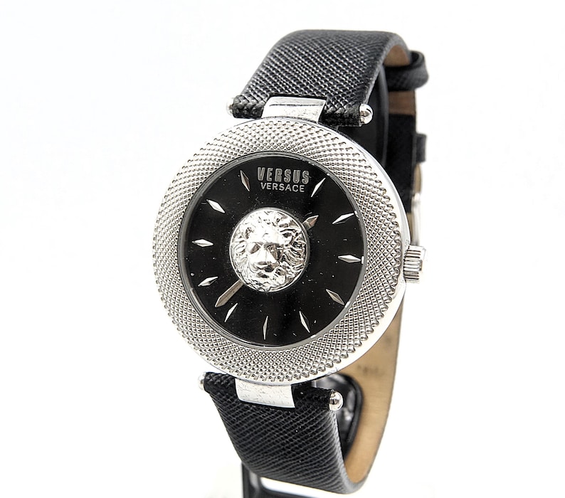 Puede incluir: Un reloj de pulsera Versus Versace con esfera negra, un emblema de cabeza de le&oacute;n plateado en el centro, marcadores de hora plateados y un bisel plateado texturizado. El reloj cuenta con una correa de cuero negro texturizado y hebilla.