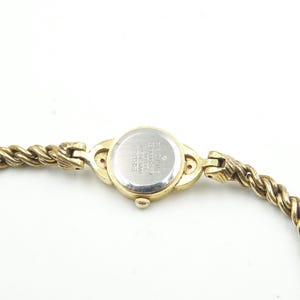 Puede incluir: Un reloj de pulsera dorado con una correa trenzada en forma de cuerda. La esfera del reloj es redonda y la parte trasera del reloj es plateada con texto. La correa se conecta a la esfera con cierres decorativos.