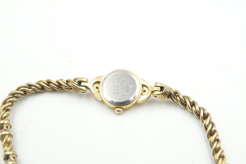 Puede incluir: Un reloj de pulsera dorado con una correa trenzada en forma de cuerda. La esfera del reloj es redonda y la parte trasera del reloj es plateada con texto. La correa se conecta a la esfera con cierres decorativos.