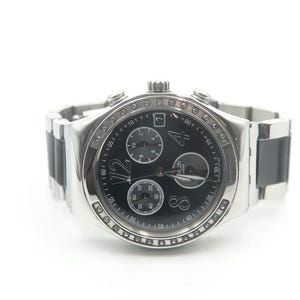 May include: A silver-tone chronograph wristwatch with a black dial, featuring three sub-dials, a date window, and large numerals at the 12, 4, and 8 o'clock positions. The bezel is accented with small clear stones, and the band has black links.