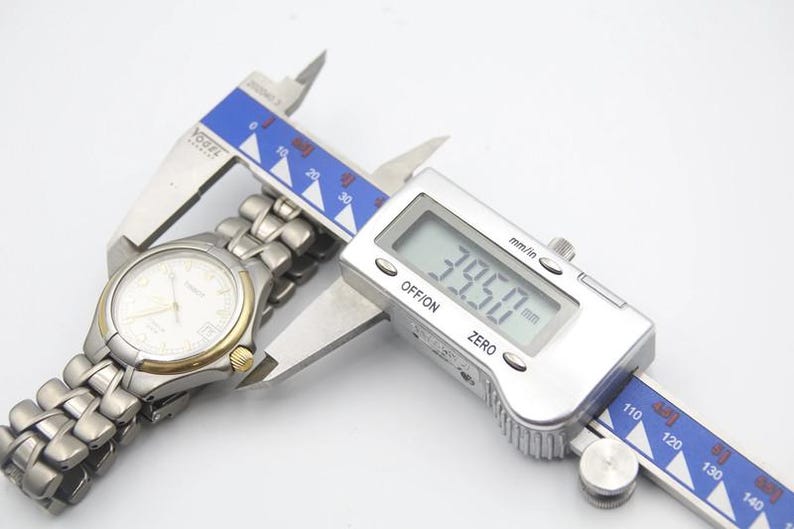 May include: A silver watch with a white face and a silver metal band is being measured with a digital caliper. The caliper reads 39.50 mm.