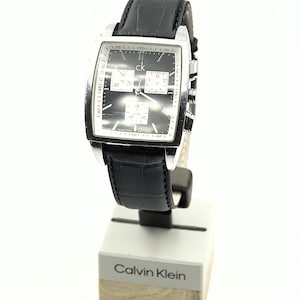 May include: A Calvin Klein wristwatch with a black face and silver-tone case. The watch features a black leather band with a crocodile pattern. The watch is displayed on a white and silver stand with the Calvin Klein logo.
