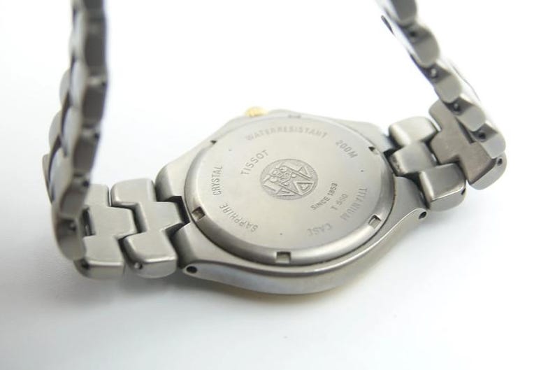 May include: Back view of a silver Tissot watch with a titanium case and sapphire crystal. The watch is water resistant to 100 meters and has a date window.
