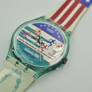 Puede incluir: Un reloj de pulsera Swatch con caja de color verde azulado translúcido y esfera con el texto Atlanta 1996 y Official Timekeeper of the Olympic Games. La esfera muestra corredores y una antorcha olímpica. La correa tiene un diseño de bandera.