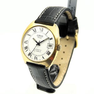 Peut inclure: Une montre automatique Oris Star dorée avec un cadran blanc, des aiguilles noires et des chiffres romains. La montre a un affichage de la date et un bracelet en cuir noir avec des coutures blanches.