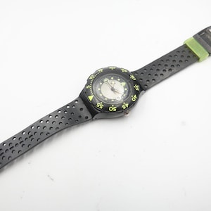 以下が含まれることがあります： 黒い腕時計、穴あきバンドと丸い文字盤。 黄色のマーキングとバンドの緑色のアクセントが付いた黒いベゼルがあります。 時計の文字盤はシルバーで、黒い針が付いています。