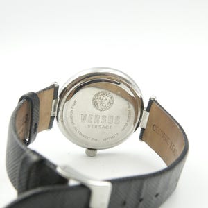 Puede incluir: Una caja de reloj de acero inoxidable plateado con una correa de cuero negro texturizado. La parte posterior del reloj presenta un emblema de cabeza de le&oacute;n y el texto VERSUS VERSACE, JAPAN MOVEMENT, ALL STAINLESS STEEL y WATER RESISTANT 30M.