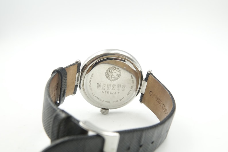 Puede incluir: Una caja de reloj de acero inoxidable plateado con una correa de cuero negro texturizado. La parte posterior del reloj presenta un emblema de cabeza de le&oacute;n y el texto VERSUS VERSACE, JAPAN MOVEMENT, ALL STAINLESS STEEL y WATER RESISTANT 30M.