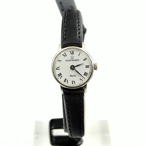 May include: A silver-toned Michel Herbelin Paris watch with a white face and black Roman numerals. The watch has a black leather band. The watch face reads "Paris" below the hands.