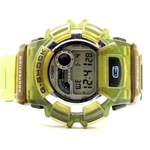 Può includere: Un orologio digitale G-Shock con cassa verde e marrone trasparente e cinturino verde chiaro. Il quadrante mostra l'ora e la data. L'orologio ha la scritta "G-Shock" sul quadrante.