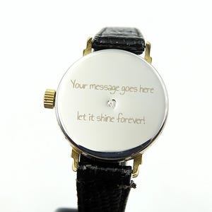 May include: Close-up of a silver watch with a black leather band. The watch face has gold accents and a small diamond. Engraved on the watch face is the text: "Your message goes here, let it shine forever!"
