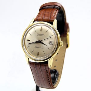 May include: A gold-toned Richard Automatic watch with a light-colored face and a brown leather band. The watch face displays the date and the word "Perfection". The band is approximately 0.75 inches wide.