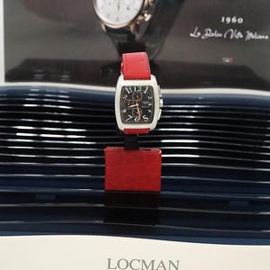 Può includere: Orologio Locman rettangolare con quadrante bianco, quadrante nero e cinturino in pelle rosso. L'orologio è esposto su un supporto rosso su sfondo scuro. Il quadrante ha numeri e lancette nere. Il marchio "LOCMAN ITALY" è visibile in basso.