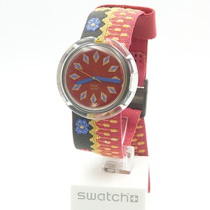 May include: A Swatch watch with a red face and a patterned strap. The watch face has diamond-shaped accents and blue hands. The strap is mainly red with yellow and black woven details. The watch is displayed on a white stand.