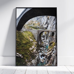 Op de afbeelding: Een landschapsfoto van een stenen brug over een kolkende rivier in een bergachtig gebied. De brug is gebogen en heeft een rustieke, verweerde uitstraling. De rivier stroomt door een smalle kloof, en de omliggende bergen zijn bedekt met weelderige groene vegetatie.