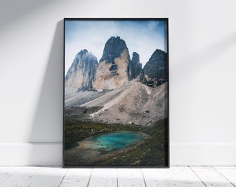 May include: A framed print featuring a scenic mountain landscape. The image showcases towering, rugged peaks with a mix of gray and tan rock formations. A turquoise lake sits at the base of the mountains, surrounded by green vegetation, under a cloudy sky.