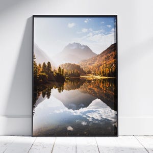 May include: A framed landscape print featuring a serene lake reflecting the surrounding mountains and autumn foliage. The image captures a sunny day with a clear blue sky, creating a peaceful and scenic view. The frame is black.