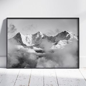 Könnte beinhalten: Schwarz-Weiß-Druck eines gerahmten Bergpanoramas. Die schneebedeckten Gipfel erheben sich über den Wolken. Der Druck wird vor einer weißen Wand und auf einem weißen Holzboden präsentiert.