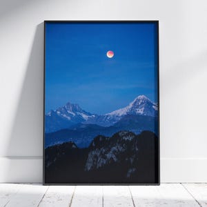 Può includere: Una stampa incorniciata con un paesaggio montano sotto un cielo blu intenso. L'immagine mostra cime innevate e una luna rosso sangue. La stampa è in una cornice nera ed è esposta contro un muro bianco.