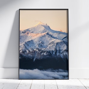 Op de afbeelding: Een ingelijste print van een besneeuwde bergketen. De bergtoppen worden verlicht door een warm, goudkleurig licht, dat contrasteert met de koele blauwe en witte tinten van de sneeuw en de lucht. De onderkant van de afbeelding toont een donker bos en een laag wolken.