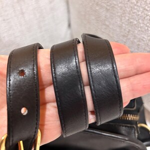 May include: Close-up of a black leather handbag with a gold-colored zipper and buckle. The bag's straps are visible, showing the leather's texture and stitching. The bag appears to be well-made and durable.
