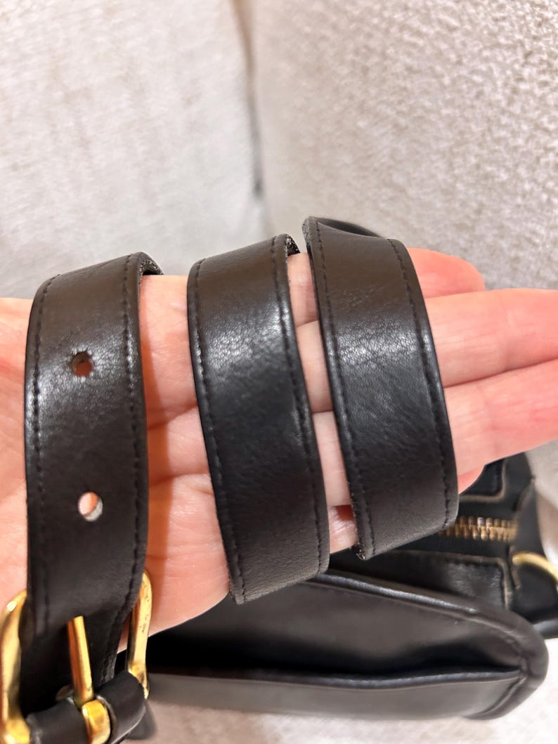 May include: Close-up of a black leather handbag with a gold-colored zipper and buckle. The bag's straps are visible, showing the leather's texture and stitching. The bag appears to be well-made and durable.