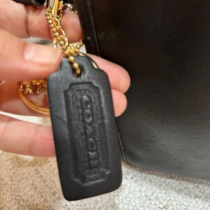 May include: Close-up of a black leather Coach handbag with a gold-tone chain and hardware. The handbag features a black leather tag with the embossed Coach logo. The bag is held by a person.