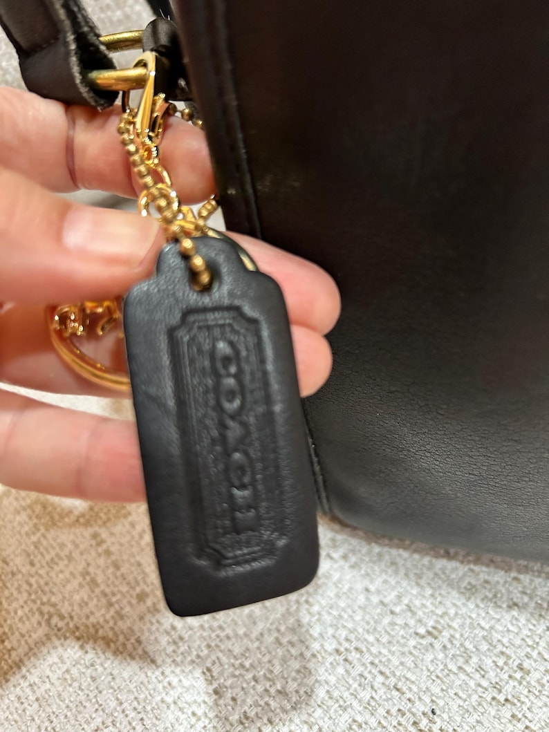 May include: Close-up of a black leather Coach handbag with a gold-tone chain and hardware. The handbag features a black leather tag with the embossed Coach logo. The bag is held by a person.